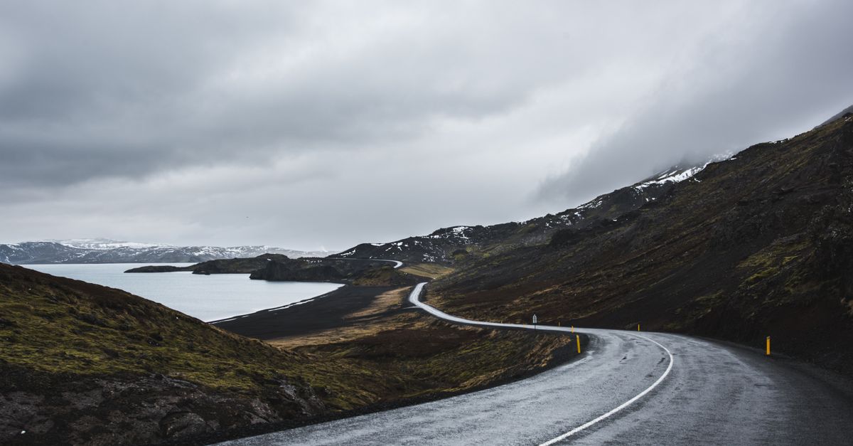A winding paved road beside water and snowy hills in Iceland