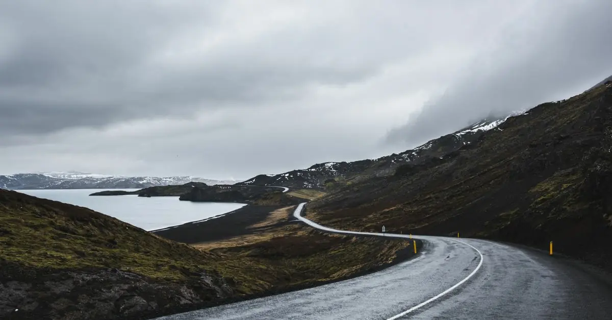 A winding paved road beside water and snowy hills in Iceland